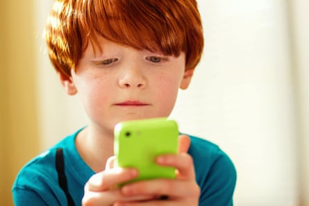 Young red-haired boy playing game on a mobile phone in a bright lime green case; he is concentrating intently as he holds the phone in both hands.