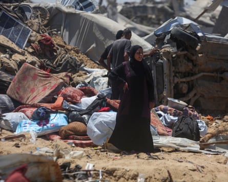 there are piles of mattresses, bedding and textiles behind her and men are looking at a wrecked, overturned vehicle.