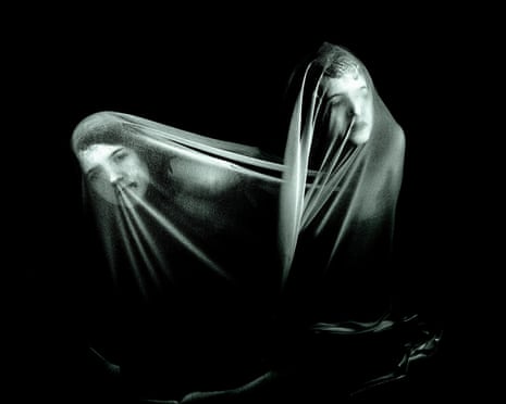 Two figures draped in translucent fabric against a black background, their faces partially visible through the material