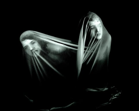 Two figures draped in translucent fabric against a black background, their faces partially visible through the material