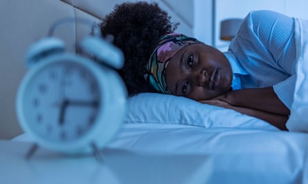 A young woman lying in bed looking anxiously at an alarm clock (in the foreground)