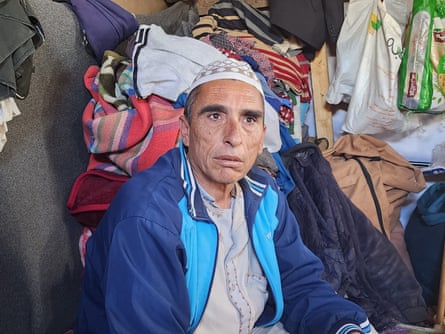 Jalal Muhammad Harb Warsh Agha sits next to various rugs and pieces of clothing in a makeshift space