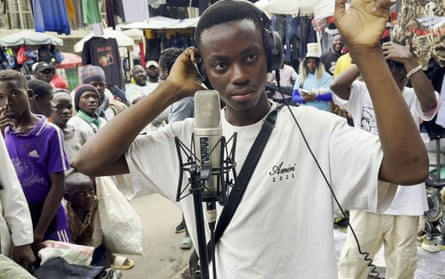 Young African man sings into microphone while wearing headphones, on the street with a crowd around him.