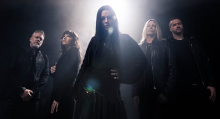 A picture of five musicians. In the centre is a woman with long black hair. Behind her are three male musicians, two with beards, one with long blond hair, and one female musician with brown hair