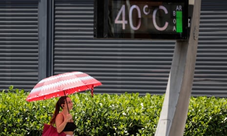 Young woman with long dark hair in ponytail, wearing glasses, crop top and holding large red umbrella, passes beneath digital sign that reads "40 degrees Celsius."