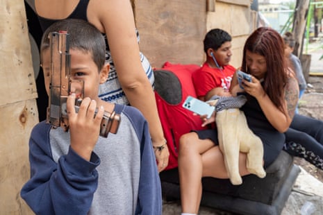 A young boy holds up a toy gun to the camera. A family with a baby sits on a bench behind him