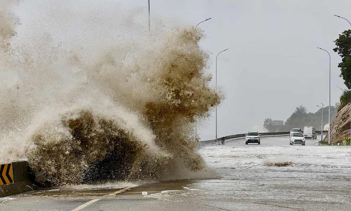 A huge ocean wave crashes on a flooding roadway with two cars driving toward it in the distance.