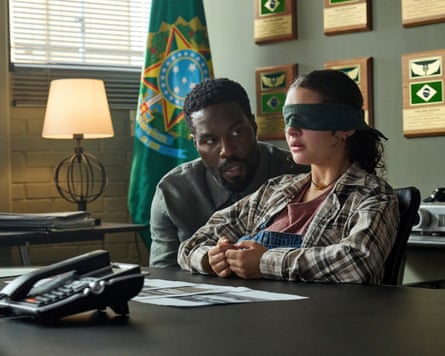 The girl sits at a desk, blindfolded, while the man speaks to her