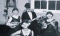 Black and white vintage photograph of four young people, three women in 30s era dresses, with a masked man at the back wearing a dinner suit. All of them are holding instruments: a guitar, ukelele, banjo or maybe a banjelele, and what could be a bouzouki.