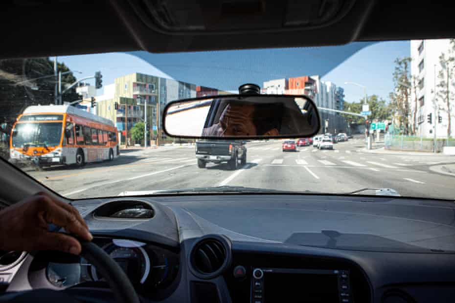 A man drives a car down a wide downtown street. In the rearview mirror is a reflection of his face