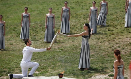 The Greek actor Mary Mina, in the role of the high priestess, passes the Olympic flame to Stefanos Douskos, who is down on one knee.