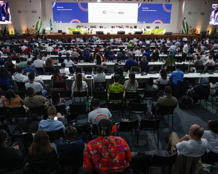 A room full of people sitting at desks, with speakers sitting on a table at the top of the room