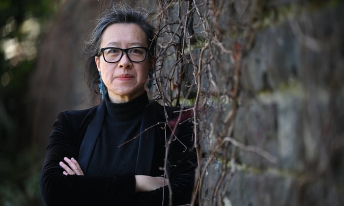 Rebecca Lim is leaning against a bluestone brick wall which is covered in some vines. She is wearing a black top and glasses, and has her arms crossed as she looks at the camera with a faint smile