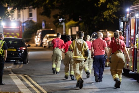 Firefighters in yellow boiler suits and dungarees and red T-shirts walk up a street, past an idling fire engine