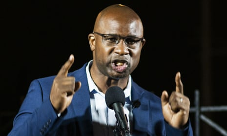 A Black man with a bald head, wire-rim glasses, blue suit and no tie raises both index fingers as he speaks into a microphone; he is well-lit and background is black, indicating he is outside at night.