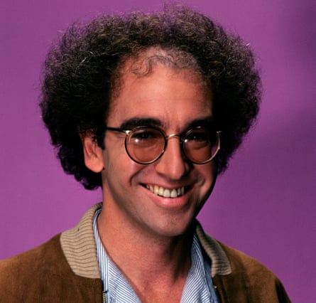 Larry David, with curly hair and round glasses, smiles broadly against a purple background
