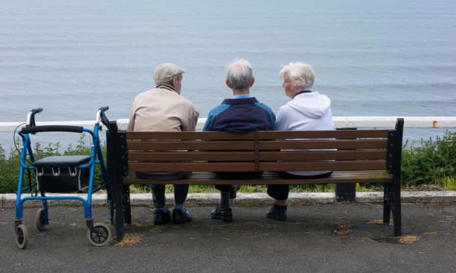 Two elderly men and an elderly woman sitting on a bench overloking sea. 