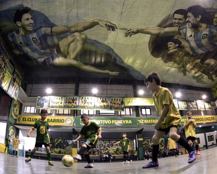 Children play football on an indoor pitch that has a mural on its ceiling of Lionel Messi and Diego Maradona in the style of Michelango’s Creation of Adam.