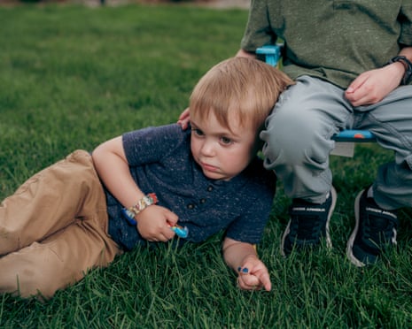A blonde toddler lies in the grass and leans against his brother sitting in a chair