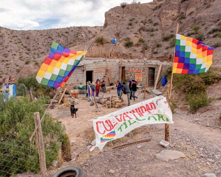 A group of people gather by low brick buildings set in a barren hillside. In front of them is a banner with an obscured slogan on it and two colourful flags on either side.