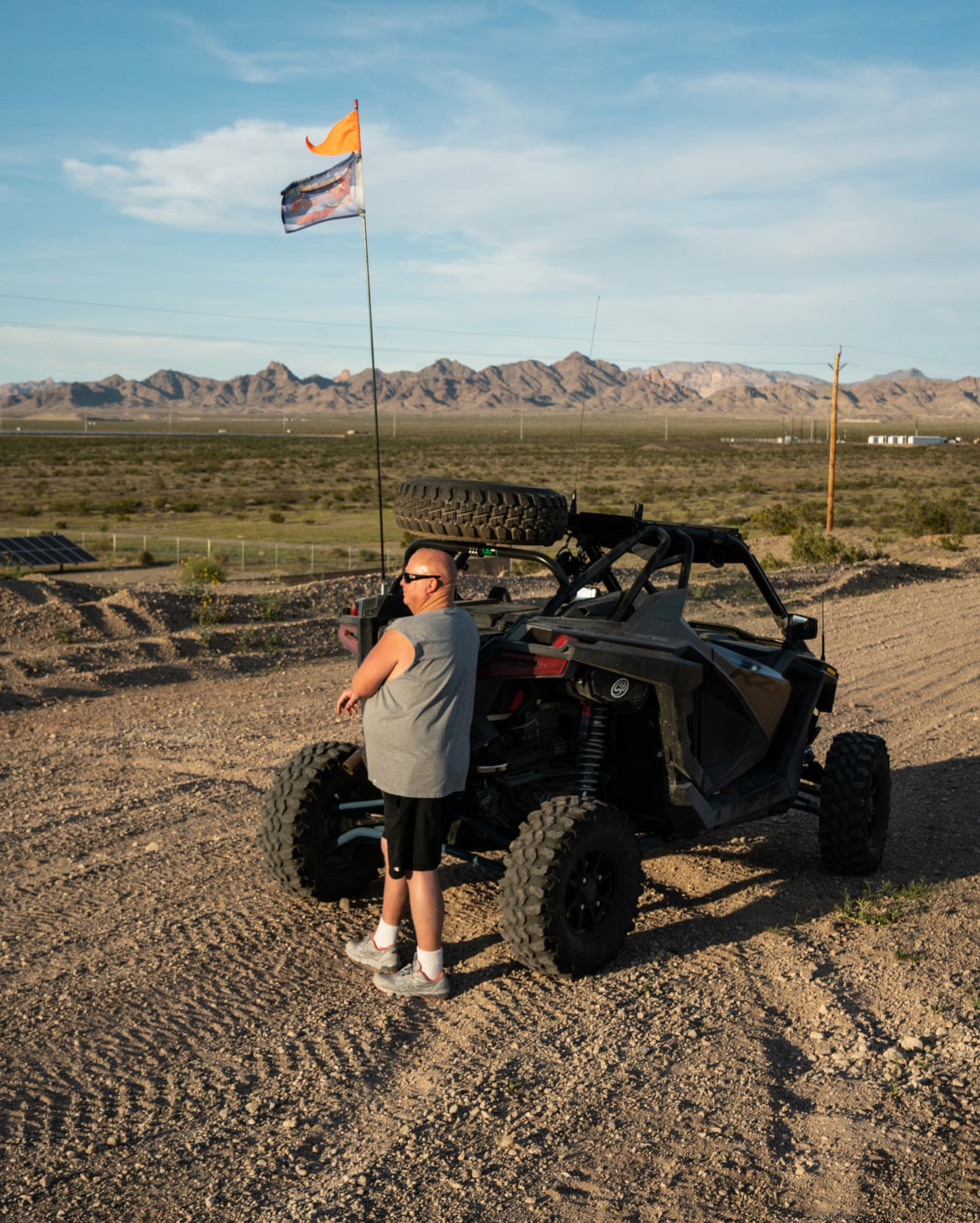 man stands next to a vehicle with a flag on it with mountains in the back