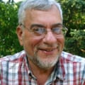 A person with short white hair and a beard who is wearing glasses poses outside