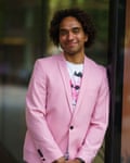 Joseph Coelho: he wears a pink jacket over a white T-shirt with a pink-faced figure on it, and is leaning in a doorway. He is quite young and has afro hair. 