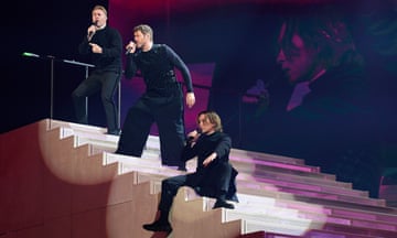 The three members of Take That dressed in black at various levels of a white staircase on stage.