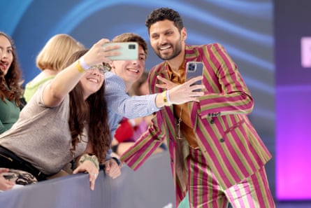 Indian man, red and brown striped blazer, smiles and poses for selfies with fans.