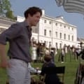 Hugh Grant wears a dark shirt and light jeans and stands in front of Kenwood House.