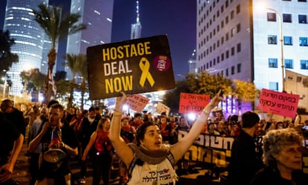 People march through the street. Tall buildings are lit up behind them. Many are carrying signs, including one that says ‘hostage deal now’.