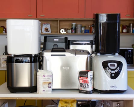 Five different bread maker machines stand on a kitchen counter beside bags of bread flour