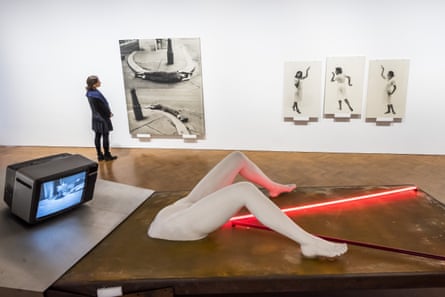 Works by Valie Export. In the foreground, Geburtenbett (Birth Bed): an outsized female abdomen with crooked legs on a mattress, red neon strip lights streaming from her vagina, and a TV transmitting a Catholic mass where the head would be. On the wall in the background hang a couple of Export's other works.