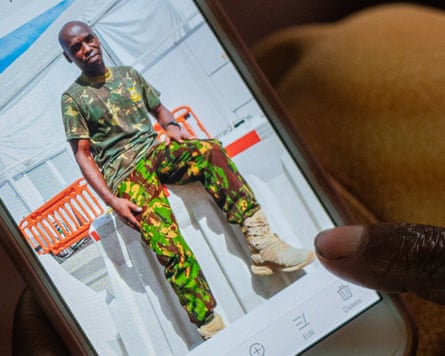 Phone screen shows a picture of Benedict Kuria wearing army combat fatigues and heavy boots; in this picture he is clean-shaven with a shaven head and he sits on a plastic road barrier.
