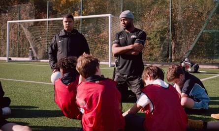 Two coaches kneel and speak to a group of pupils sitting on a football pitch near a goal