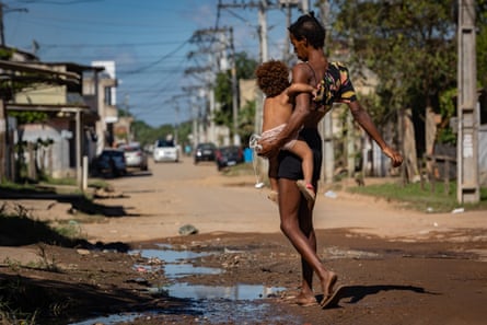 A thin woman in shorts and flip-flops with a baby on her hip crosses a dirt road in a shabby town