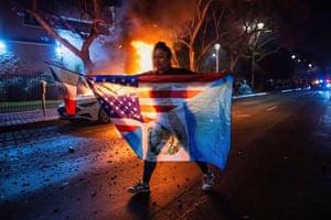 A night shot of woman carrying a flag in her outstretched arms as she walks past a burning car in the background