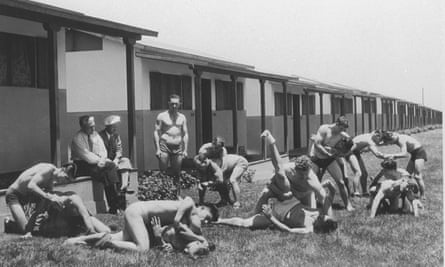 Black and white photo: a row of simple single storey huts – a lot like a 1950s UK holiday resort – in the sunshine 7 pairs of well muscled shirtless men wrestle together, while another shirtless man looks on, and two fully dressed men watch from the shade of a hut.