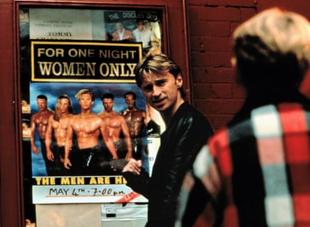 Robert Carlyle points at a poster of bare-chested men while another man looks on.