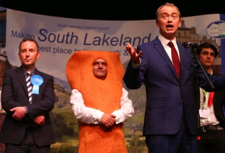 Man dressed as a fish finger stands behind Tim Farron at a vote count