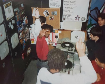 In a 1990s record shop, young men gather around decks, making peace signs and raised fists