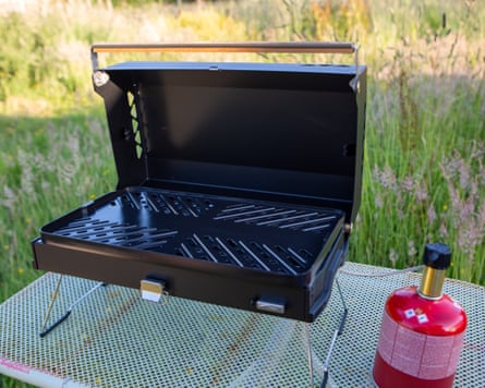 The Primus Kuchoma Portable Grill attached to a small red gas canister. Both are on a metal camping table in a field