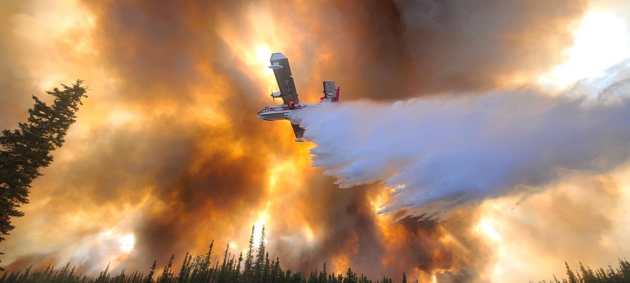 An aircraft soars above the trees, through a sky filled with smoke and orange from flames, as it drops a load of water.