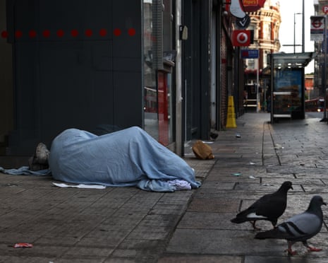 A person sleeps under a blue blanket on a pavement in a doorway, with two pigeons nearby