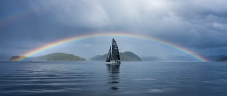 A yacht with a black sail in a spectacular still waterway perfectly framed by a magnificent rainbow.