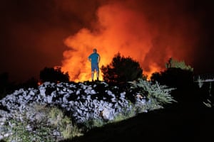 A man in blue T-shirt and shorts stands on a mould of rock as bushes as he looks towards a wildfire