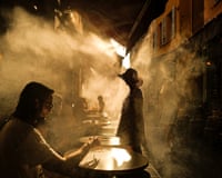 A person in a hat stands in a misty, sunlit alley near a coffee shop