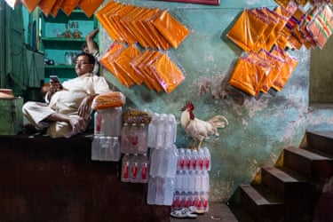 A merchant relaxes in his shop near stacked water bottles as a rooster perches nearby