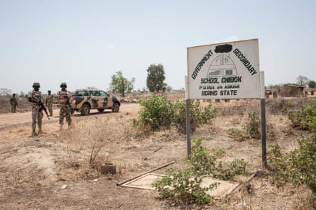 Four soldiers in camouflage and helmets stand near their military pickup truck behind a school sign in a scrubby desert-like landscape