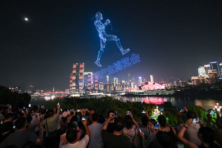 Night-time scene as people watch a huge 3D image of a man running and Chinese lettering appear in the sky above a river, with lit-up skyscrapers in the background
